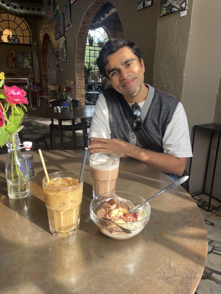Picture of a 20-year-old man in a cafe with drinks and food in front of him.